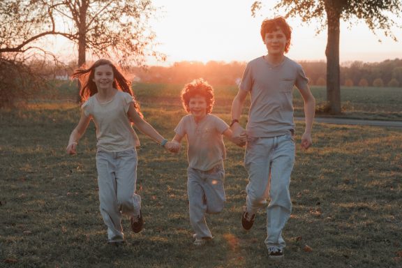 Lebendige Geschwisterfotografie Drei Kinder laufen gemeinsam über eine Wiese im warmen Abendlicht.