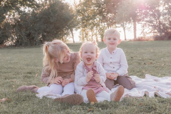 Fröhliche Geschwisterbilder Drei kleine Kinder sitzen nebeneinander auf einer Decke im Gras und lachen.
