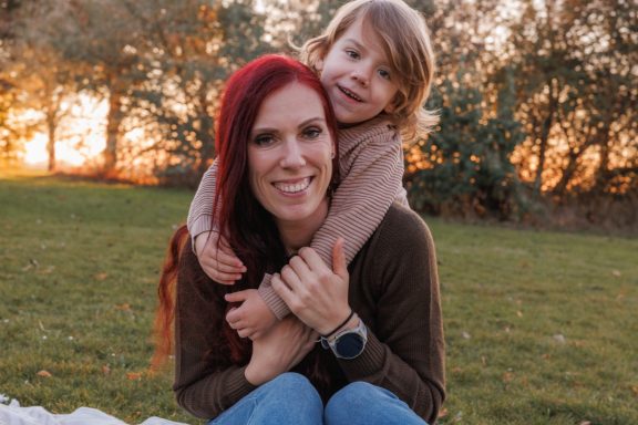 Liebevolle Mama-Kind-Momente Lächelnde Mutter mit roten Haaren trägt ihre Tochter auf dem Rücken und beide kuscheln miteinander.