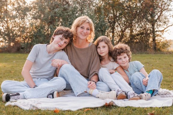 Familienfotografie im Grünen Mutter sitzt mit ihren drei Kindern eng aneinandergekuschelt auf einer Decke im Grünen.